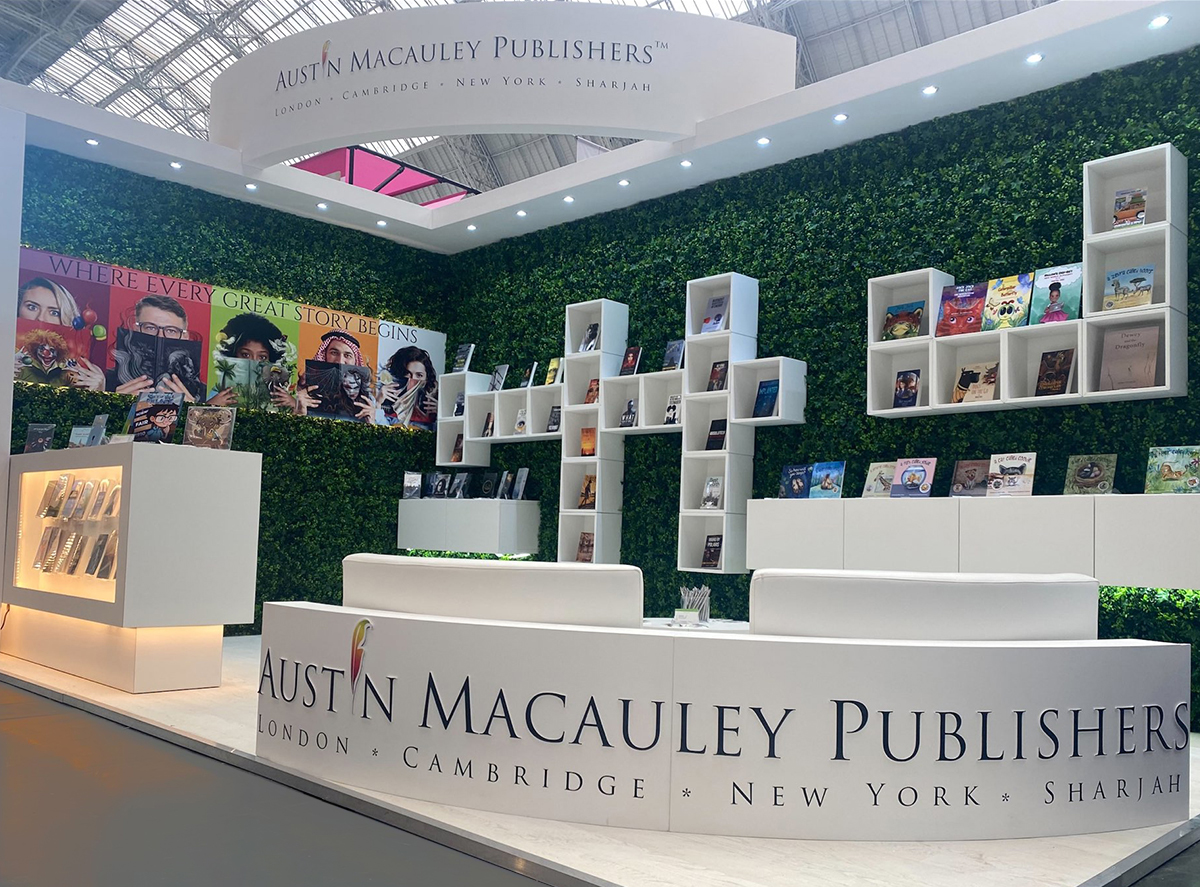 Image shows a photo of an Austin Macauley Publishers stand at a book fair. On a green leafy wall there are a series of white box bookshelves. There is also a white curved sign with the logo of Austin Macauley Publishers.