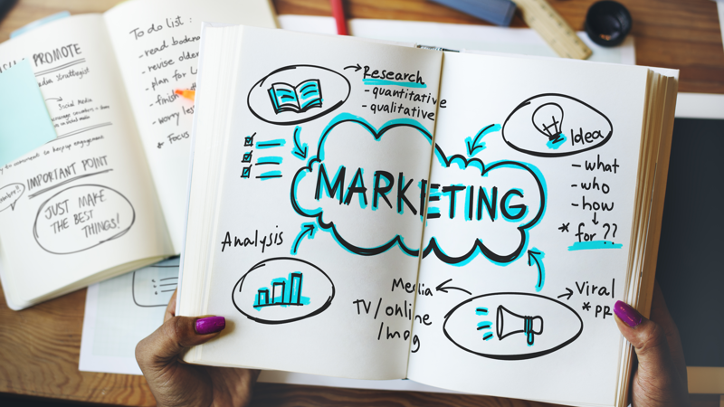 Image shows someone holding open a book with the words 'Marketing' written in the middle and images of a book, graph, megaphone and a bulb surrounding the word. In the background, there are other books open.