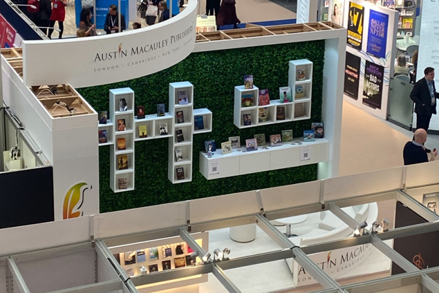 Image shows a zoomed out photo of an Austin Macauley Publishers stand at a book fair. On a green leafy wall there are a series of white box bookshelves. There is also a white curved sign with the logo of Austin Macauley Publishers.