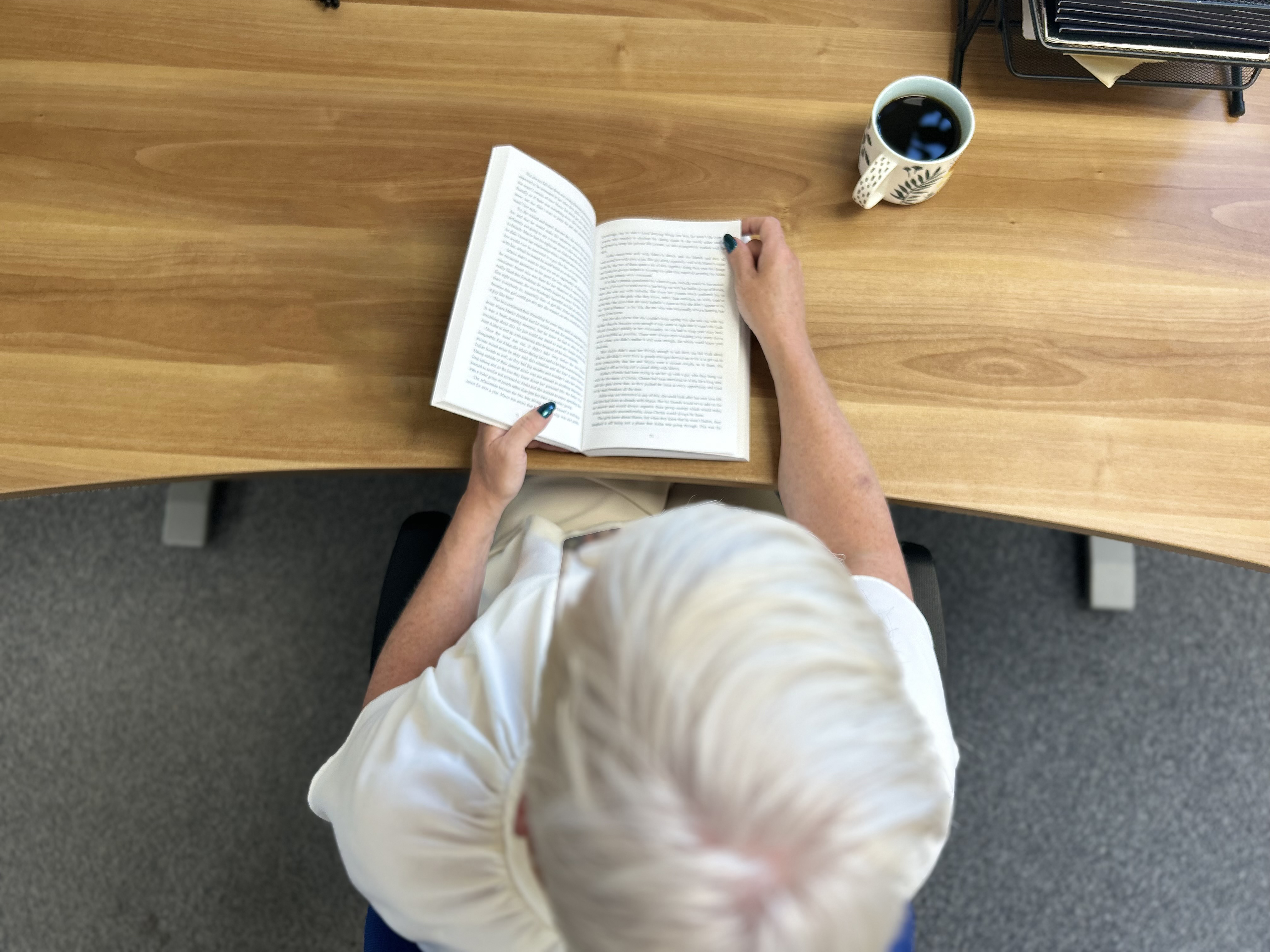 Image shows an aerial view of a blonde haired women reading a book sat a desk, with a mug of coffee next to her.